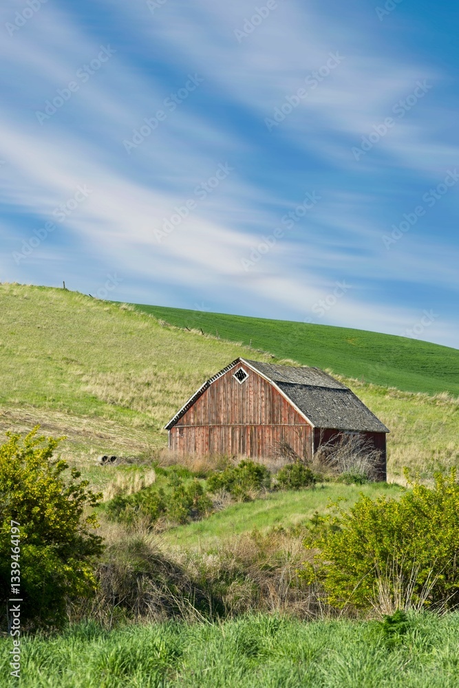 Obraz premium Rustic red barn in lush green hills under blue sky.