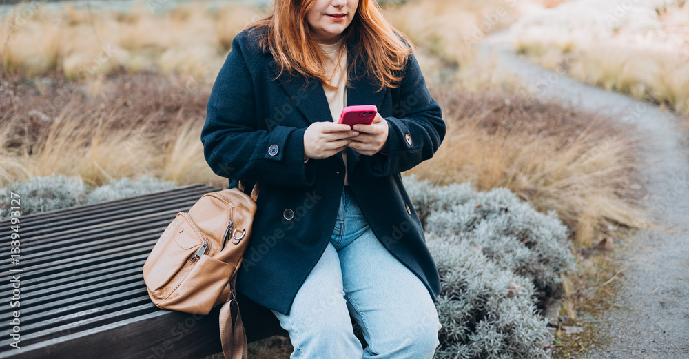 Happy woman sitting on the bench and chatting on phone outdoors. Urban, people concept. Smiling 30s redhead woman text messaging on smart phone at sunny day on the city street, spring time