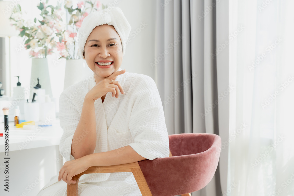 Asian middle-aged woman wearing white bathrobe and towel on head sitting indoors. Smiling with radiant skin in a bright modern room. Beauty, self-care, and wellness moment for healthy aging Asian middle-aged woman wearing white bathrobe and towel on head sitting indoors. Smiling with radiant skin in a bright modern room. Beauty, self-care, and wellness moment for healthy aging