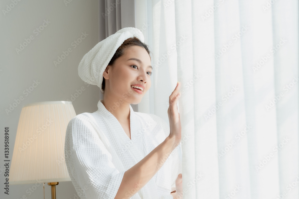 Radiant Asian woman in white bathrobe with towel on head smiling while looking through window in bright cozy bedroom. Concept of morning self care, fresh start, relaxation, and beauty wellness. Radiant Asian woman in white bathrobe with towel on head smiling while looking through window in bright cozy bedroom. Concept of morning self care, fresh start, relaxation, and beauty wellness.