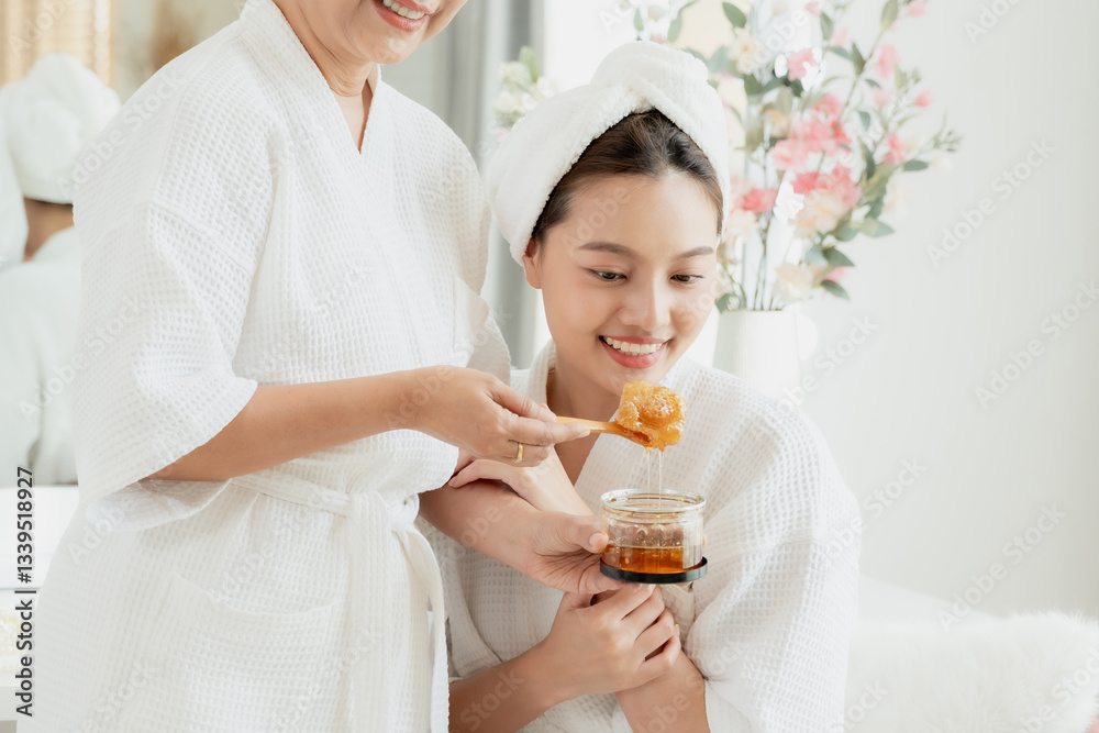 Elderly Asian woman and young adult daughter wearing white bathrobes and towels smiling while holding honey for skincare indoors. Enjoying a natural beauty treatment together. Self-care, wellness, Elderly Asian woman and young adult daughter wearing white bathrobes and towels smiling while holding honey for skincare indoors. Enjoying a natural beauty treatment together. Self-care, wellness,