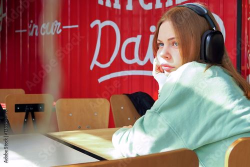 A young woman sits alone at a table in a cafe looking sad and bored, listens to music in headphones