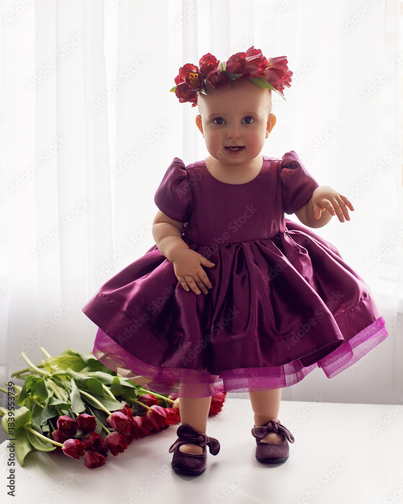 A portrait of a cheerful, beautiful one-year-old girl stands in a burgundy dress at the window of the house. He wears a wreath of flowers on his head