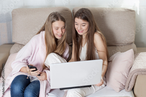 Happy young freelancers working at home using modern portable device, cheerful students in casual clothes with long hair browsing the internet studying with laptop computer in cozy home interior