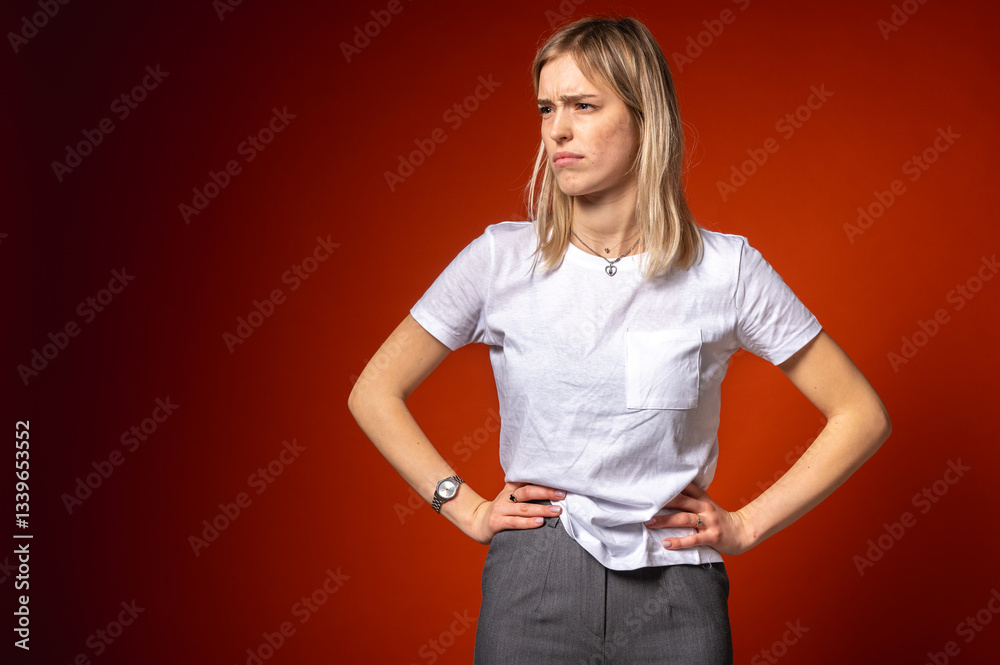 Young woman in white t-shirt making disgusted facial expression, surprised, holding hands on hips on red background. Concept of dissatisfaction, disappointment or irritation, stress