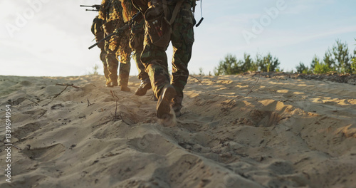 Military troops march across the sandy field, their boots kicking up dust, patrolling the area with weapons in hand, fully prepared for a tactical exercise.