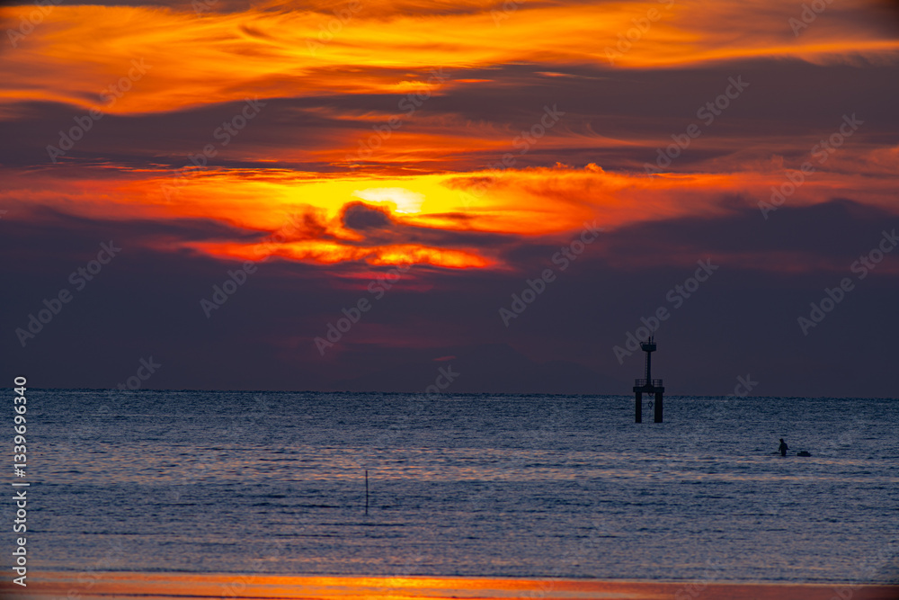 Fototapeta premium A striking seascape capturing the vivid hues of a sunset sky blending orange, red, and purple over the calm ocean. A navigation tower stands against the horizon, adding depth and dramatic scene. .