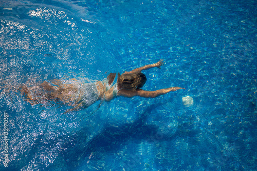 A young girl in a swimsuit swims underwater in a pool with a blue bottom. The girl dived