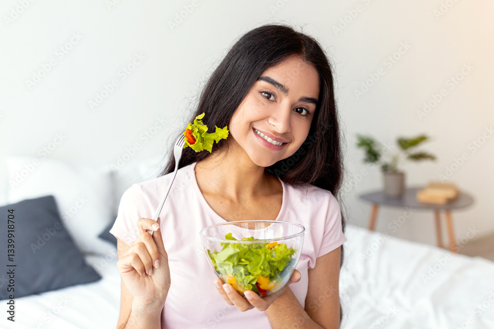 A young Indian woman smiles while holding a bowl of fresh salad in her hand. She is wearing a casual outfit and looks fit, embracing a healthy lifestyle at home. A young Indian woman smiles while holding a bowl of fresh salad in her hand. She is wearing a casual outfit and looks fit, embracing a healthy lifestyle at home.