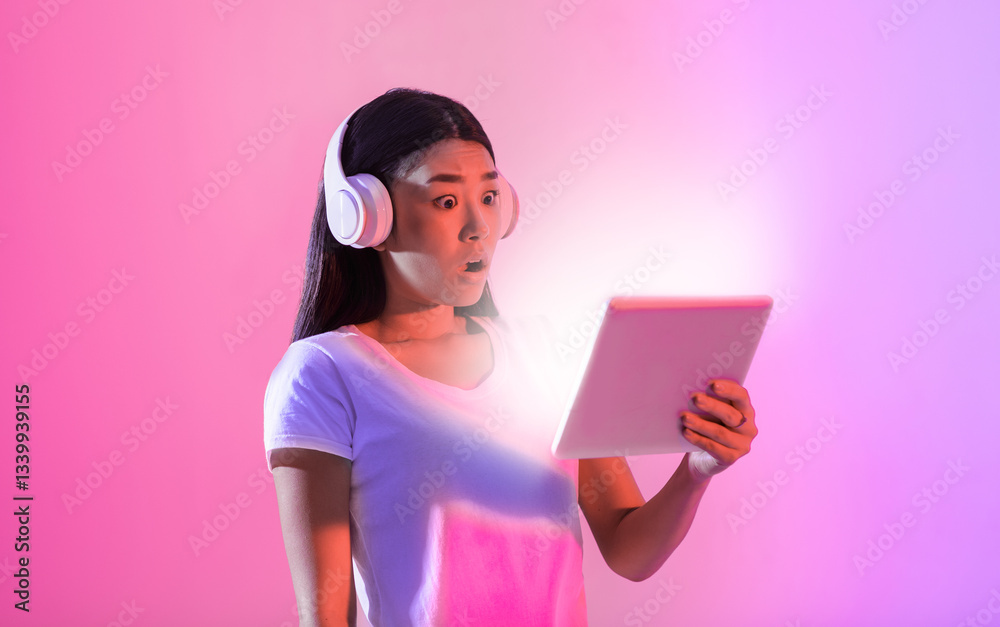 Asian woman wearing headphones expresses amazement as she looks at a tablet. The vibrant pink and purple background adds to the energetic atmosphere of her reaction. Asian woman wearing headphones expresses amazement as she looks at a tablet. The vibrant pink and purple background adds to the energetic atmosphere of her reaction.