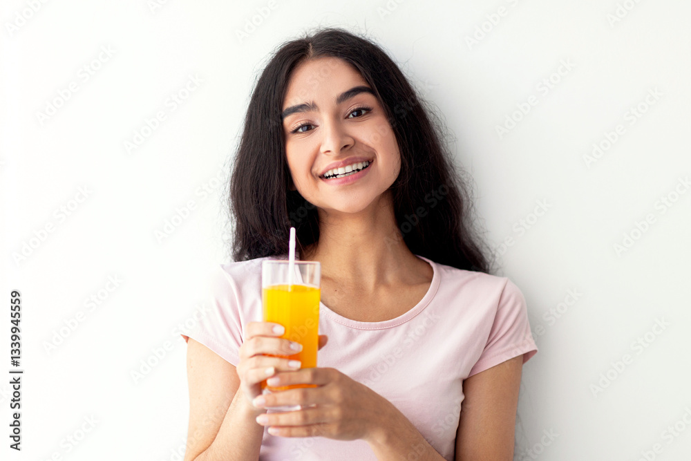 A young Indian woman with long hair smiles while holding a glass of orange juice. She is dressed in a casual t-shirt and appears to be enjoying a refreshing drink at home. A young Indian woman with long hair smiles while holding a glass of orange juice. She is dressed in a casual t-shirt and appears to be enjoying a refreshing drink at home.