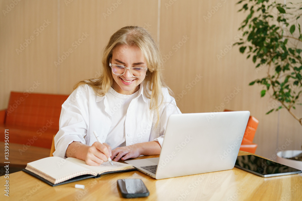 Young woman writing in notebook while working on laptop in bright, modern workspace during daytime Young woman writing in notebook while working on laptop in bright, modern workspace during daytime
