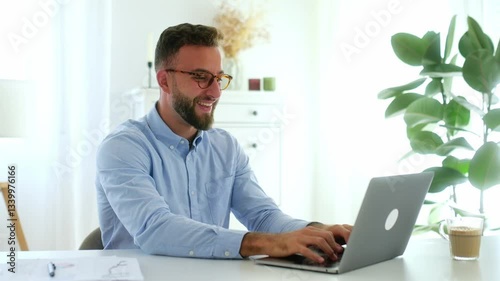 Achievement goal. Cheerful successful businessman with glasses in a blue shirt, raising his fists while looking at his laptop screen, celebrating a success in a bright modern office, smiling happily