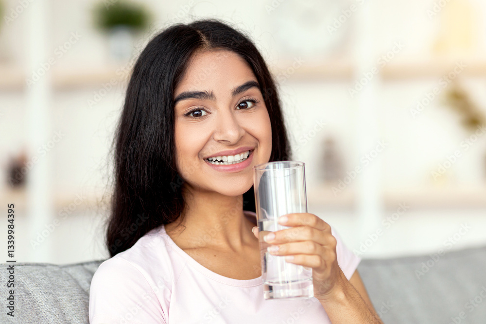 A millennial Indian woman smiles brightly while holding a glass of water at home, promoting a healthy lifestyle. She embraces fitness and wellness as part of her daily routine. A millennial Indian woman smiles brightly while holding a glass of water at home, promoting a healthy lifestyle. She embraces fitness and wellness as part of her daily routine.