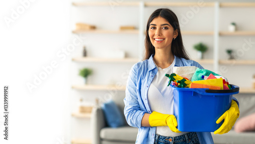 A young woman is happily holding a blue container filled with cleaning supplies while smiling. She is dressed casually and appears to be cleaning her home space during the day, copy space