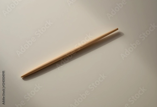 A single wooden chopstick rests diagonally on a smooth, neutral surface. Soft lighting highlights its texture and clean edges, creating a minimalist, balanced composition.