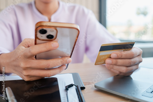 Person holding smartphone and credit card, engaging in online shopping at a home office desk with a laptop.
