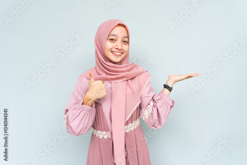 Asian Muslim young woman wearing headscarf veil hijab smiling cheerfully while looking and pointing to copy space beside her with thumbs up gesture, standing on isolated gray background.