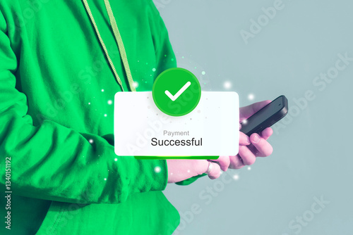 Man wearing a green hoodie holds a smartphone displaying a Payment Successful notification with a green checkmark. Online payment concept.