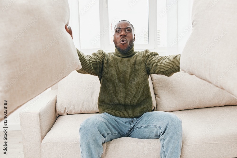Young African American man expressing frustration on a couch while holding pillows, wearing a green sweater, casual jeans, in a bright modern interior Young African American man expressing frustration on a couch while holding pillows, wearing a green sweater, casual jeans, in a bright modern interior