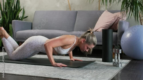 Young beautiful blonde doing knee push-ups on yoga mat in cozy living room. Gray sofa and houseplants, palm trees, dracaenas in the background. Active lifestyle concept.