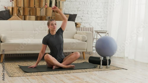 Young fit woman doing yoga, sitting on a mat with crossed legs and bending to the sides. In the background is a white sofa and a shelf with books. Active lifestyle.