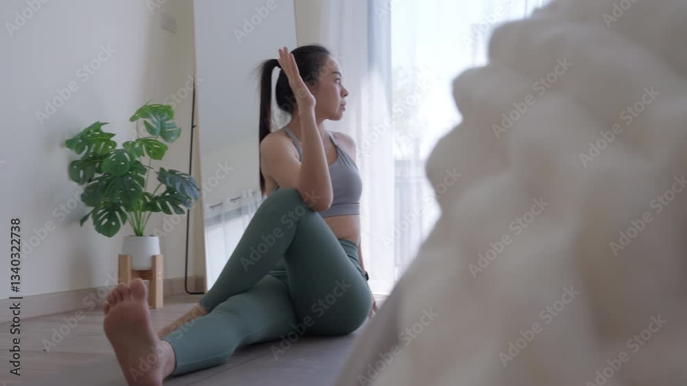 A woman is sitting on the floor in a room with a white bed in the background. She is wearing a green top and blue pants