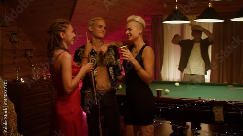Cozy wooden party: a woman in a black dress with short light hair embraces a smiling man, hand on shoulder and cup in hand, while a red-dressed friend with dreads smiles by the billiard table.
