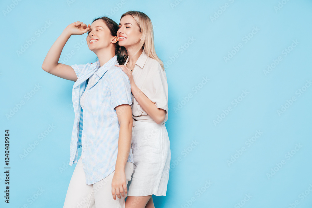 Naklejka premium Two young beautiful smiling brunette and blond hipster female in trendy summer clothes. Lovely women posing on blue background in studio. Positive models having fun. Cheerful and happy. isolated