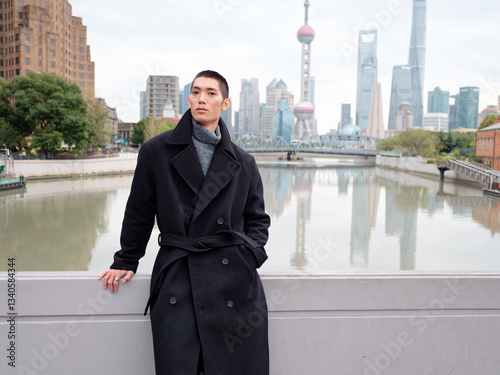 Portrait of handsome Chinese young man wearing black overcoat posing in the street, young guy with black short hair with Shanghai bund background. Male fashion, cool Asian young man lifestyle.