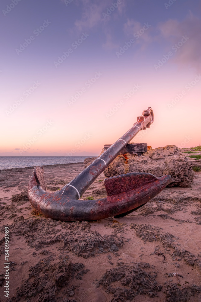 A weathered anchor rests on a rocky shore at sunset, capturing the essence of time and the sea. Perfect for travel, nature, and nautical themes. In Punta Prima beach in Minorca Island.
