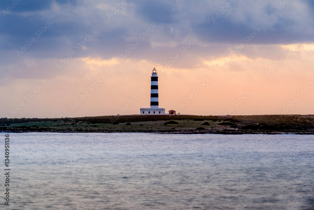 Capture the iconic beauty of Minorca with this image of a black and white lighthouse in the distance, set against twilight skies and a serene sea. Perfect for travel, nature, and scenic themes