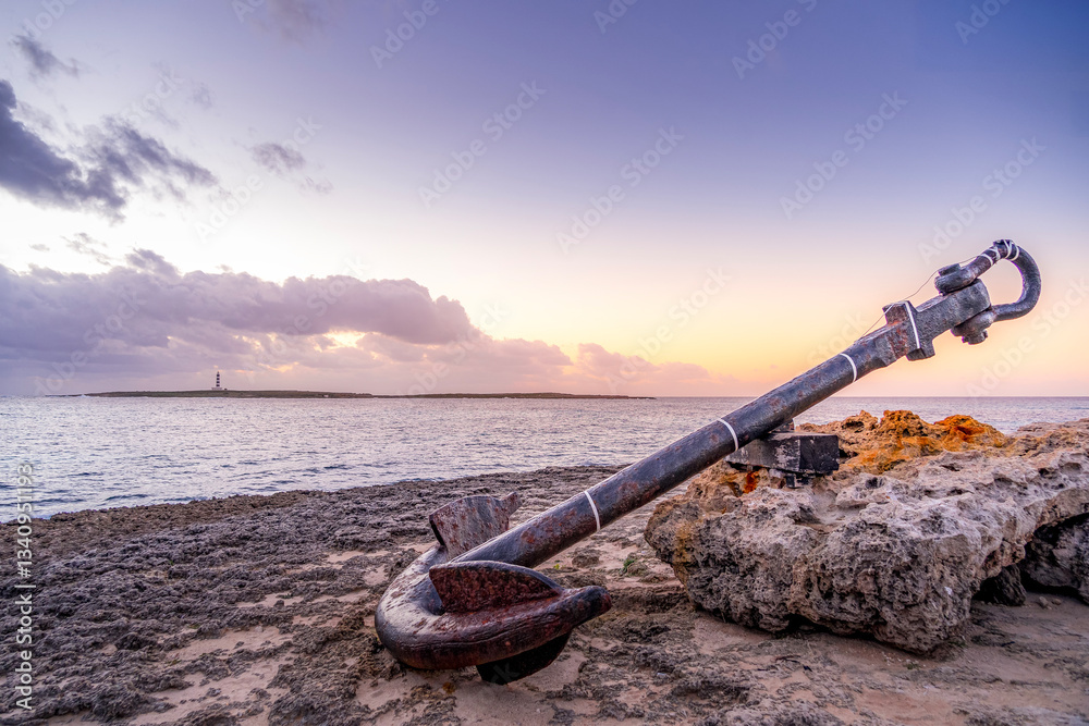 A weathered anchor rests on a rocky shore at sunset, capturing the essence of time and the sea. Perfect for travel, nature, and nautical themes. In Punta Prima beach in Minorca Island.