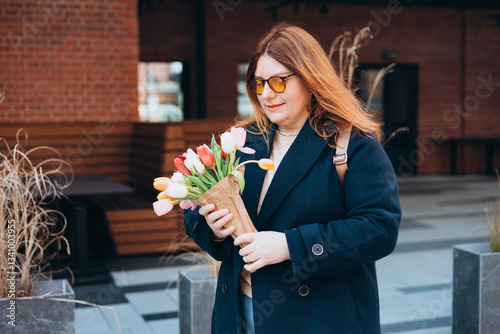 Obraz na płótnie Beautiful young woman with spring tulips flowers bouquet walking at city street. Happy girl posing outdoors. Spring portrait of pretty female on urban background. High quality photo. w salonie