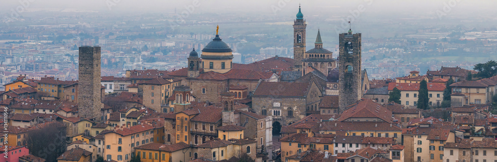 Fototapeta premium View of Bergamo's old town during sunset, featuring the Basilica of Santa Maria Maggiore, the Campanone, and medieval stone towers amid terracotta roofs.