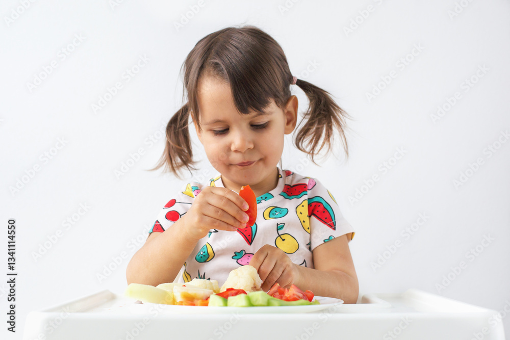 Little girl 2-3 year with pigtails sitting in high chair, carefully picking up piece of red bell pepper from plate of fresh vegetables. Healthy eating, childhood nutrition, and mindful eating Little girl 2-3 year with pigtails sitting in high chair, carefully picking up piece of red bell pepper from plate of fresh vegetables. Healthy eating, childhood nutrition, and mindful eating