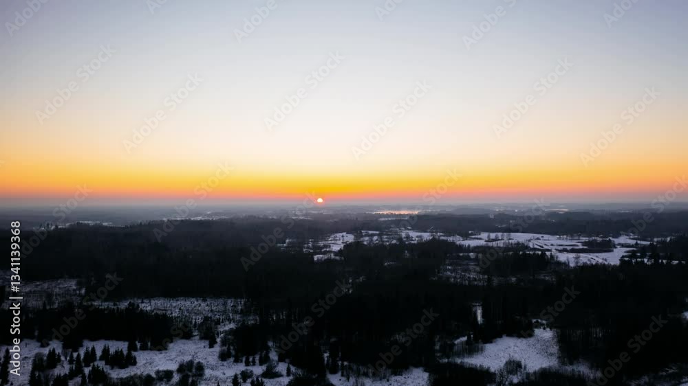 Aerial sunset hyperlapse against the sun in countryside on a cold winter evening over the forest. Advance timelapse of sun setting below horizon.