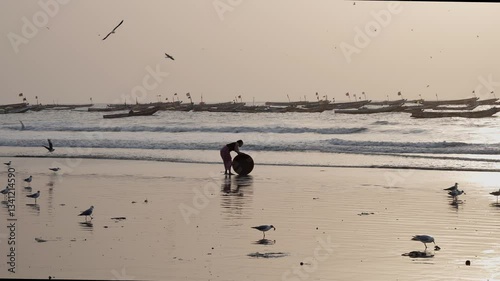 West Aferican woman washing a large bucket on a fishing beach with birds at dusk