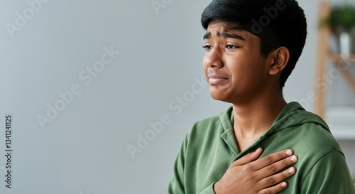Asian teen boy in green hoodie expressing sadness with hand on heart