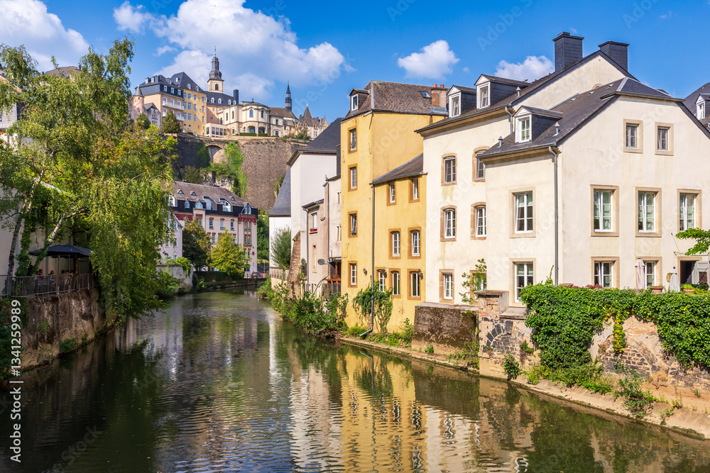 Fototapeta premium View from the Grund bridge of the Alzette river in Luxembourg City, Luxembourg, lined with old townhouses and overlooked by the old town district of Ville-Haute on a sunny summer day.