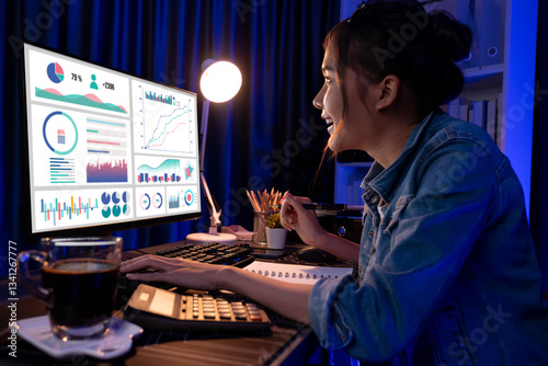 Smiling young beautiful asian creative looking on pc of profit of sales target report and writing on notebook for memo with planning invest new strategy market project at neon light night. Stratagem.