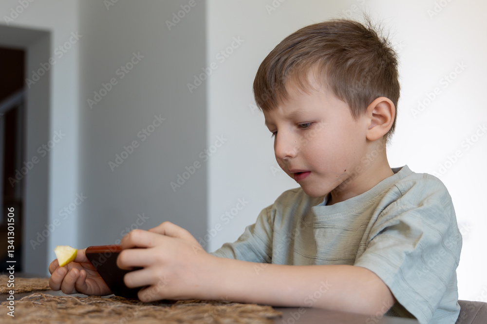 Young boy using smartphone while eating fruit. A young boy sits at a table, focused on his smartphone while holding a piece of fruit. A modern childhood moment at home. Young boy using smartphone while eating fruit. A young boy sits at a table, focused on his smartphone while holding a piece of fruit. A modern childhood moment at home.