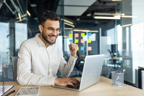 Man celebrating victory and successful achievement results at workplace. Businessman holding hand up happy smiling triumph gesture. Worker inside office working with laptop.