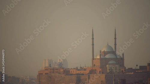 The Mosque of Muhammad Ali stands atop Cairo Citadel, its domes and towering minarets illuminated by soft haze. Modern high-rises loom in the distance, contrasting the historic fortress below.