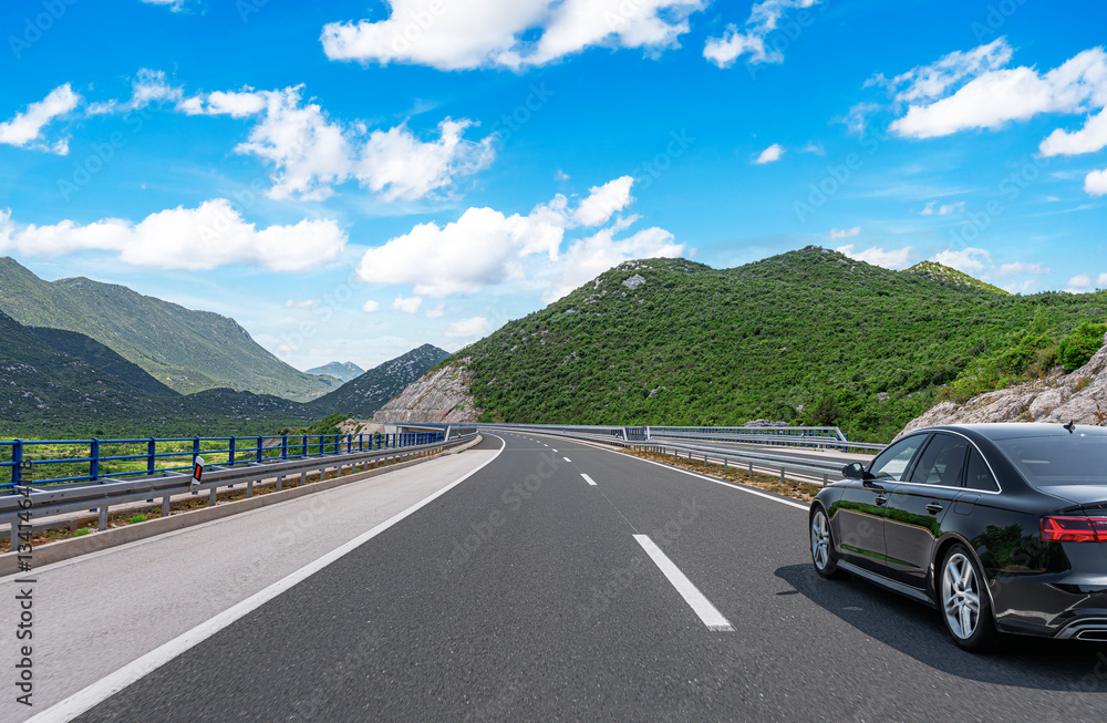 A sleek black sedan drives along a well-maintained highway winding through lush green mountains under a bright blue sky with scattered clouds.