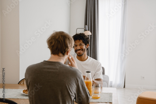 A view from the back of a young white-skinned man with short brown hair, as he sits across from a young Black man at a wooden table, in a white room. The Black man smiles widely while looking at him