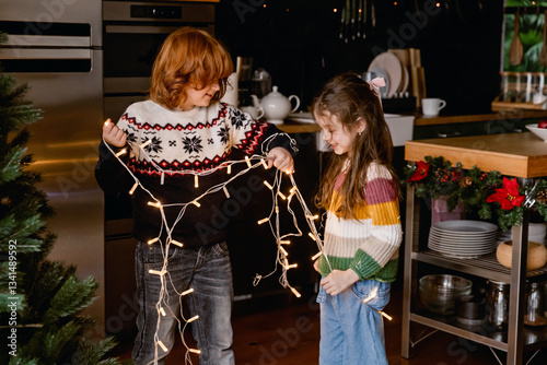 Fotografía A smiling school-aged red-haired White boy and a school-aged girl decorating a Christmas tree with string lights in a cozy festive kitchen
