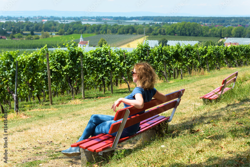 Woman relaxing on red bench overlooking scenic vineyard landscape on a sunny day
