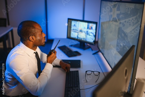 African American security officer or dispatch uses computer and walkie talkie, looks at screens