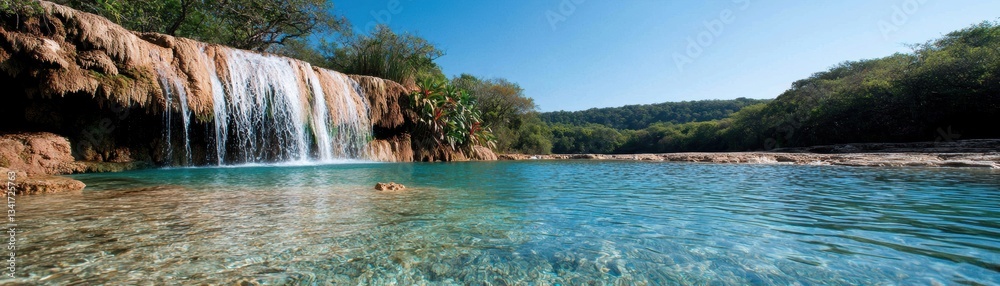 Fototapeta premium A serene waterfall cascades into a clear, tranquil pool surrounded by lush greenery under a bright blue sky.
